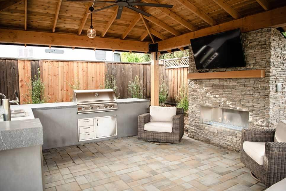 K&D Landscaping covered outdoor kitchen and lounge pavilion beside a teak dining set on a paver patio