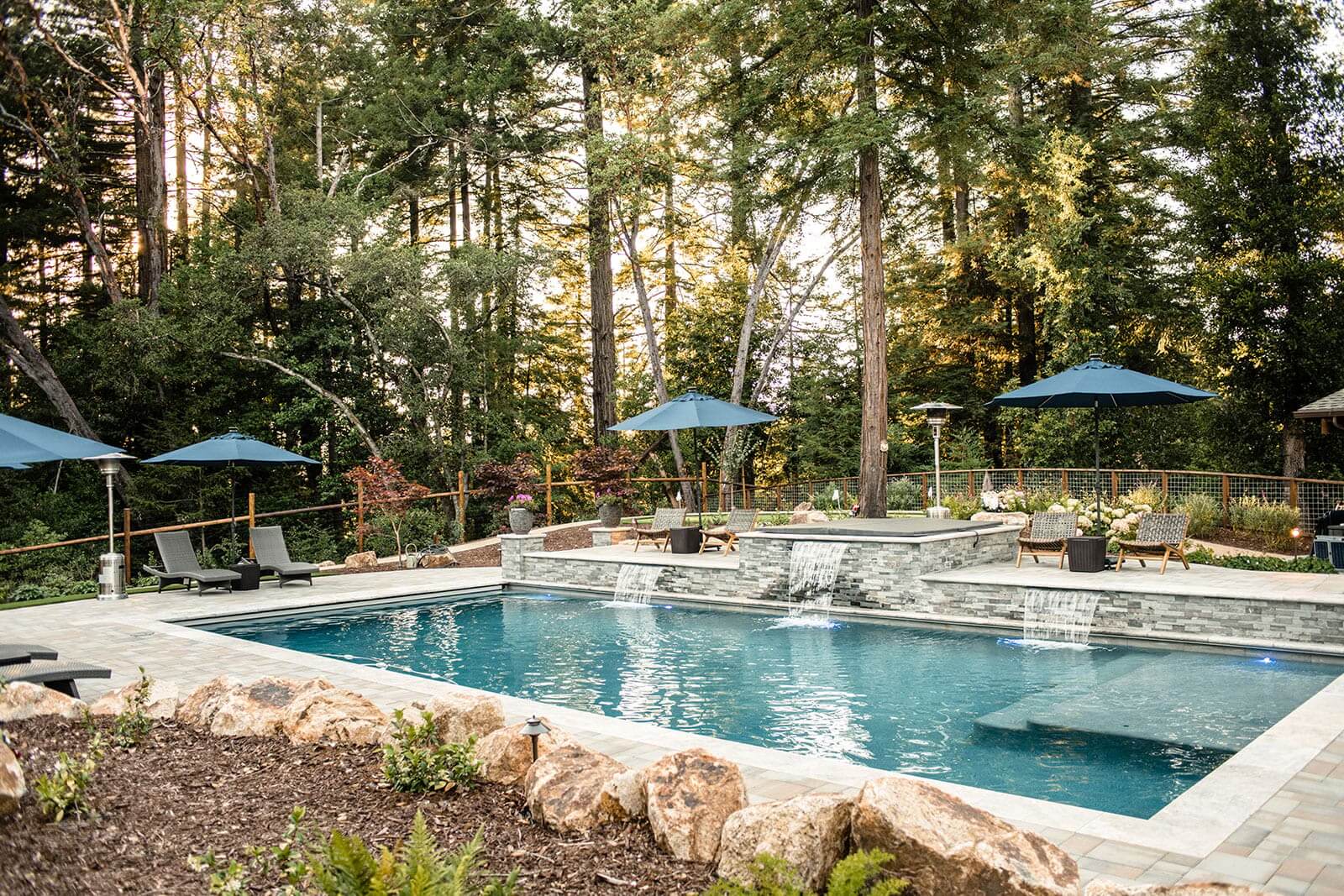 K&D Landscaping pool with three sheet waterfalls off a raised stacked-stone spa wall among redwoods