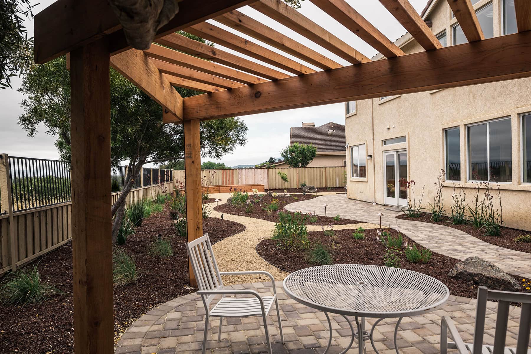 Outdoor patio area with wooden pergola, paver surface, metal furniture, and native landscape plantings completed by K&D Landscaping