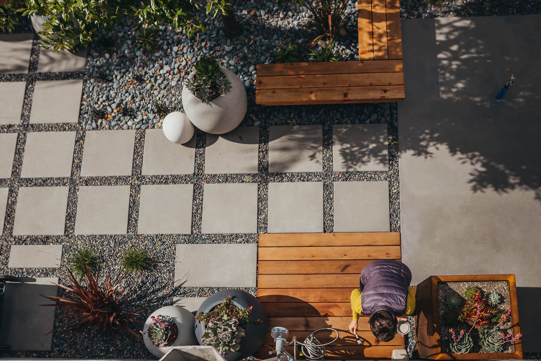 Overhead view of residential landscape with concrete pavers and gravel groundcover, white sphere planters, tall ornamental planting, redwood seating, and modern patio design by K&D Landscaping
