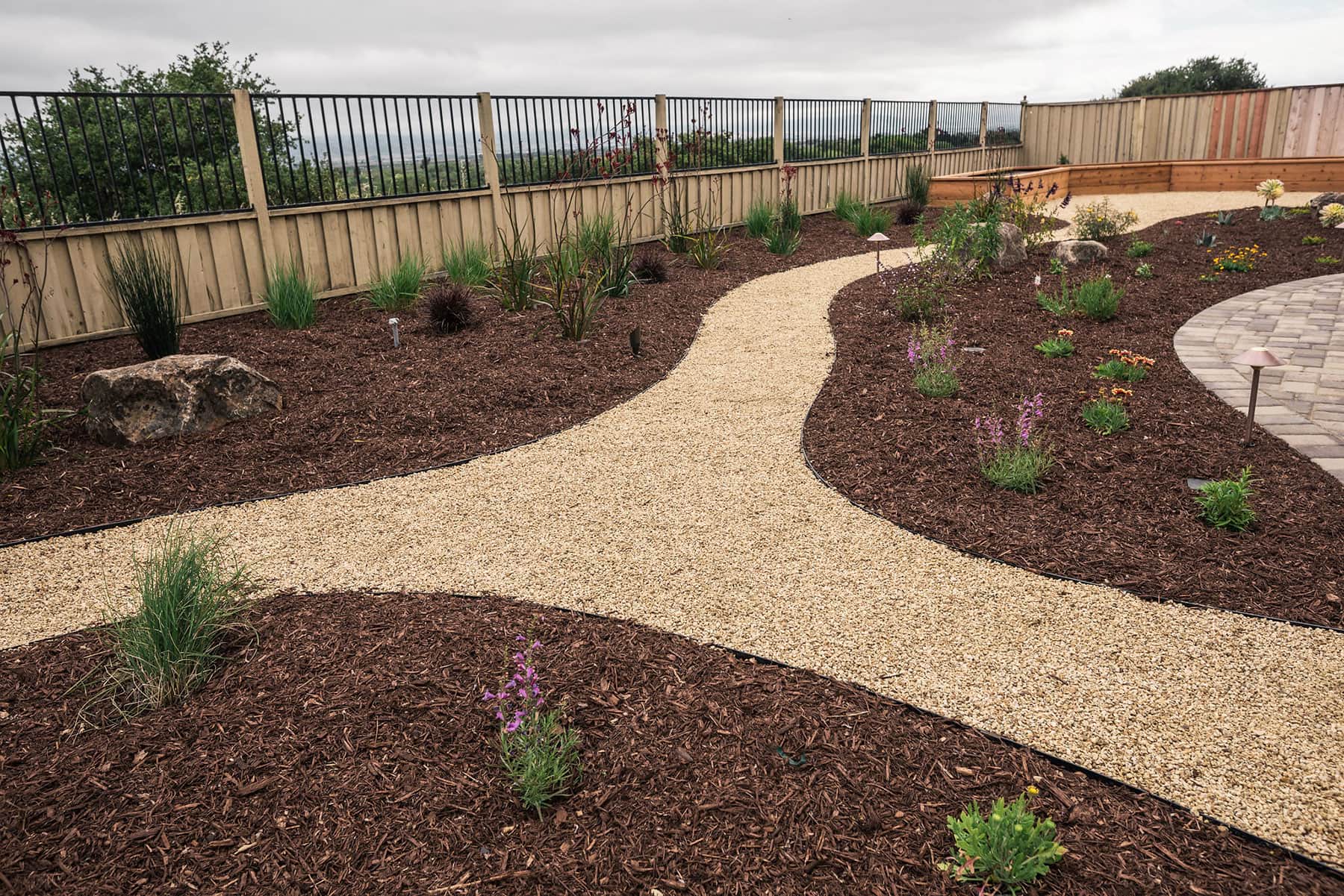 Backyard landscape design with gravel pathway curving through mulch beds, ornamental grasses, boulders, and pergola structure by K&D Landscaping