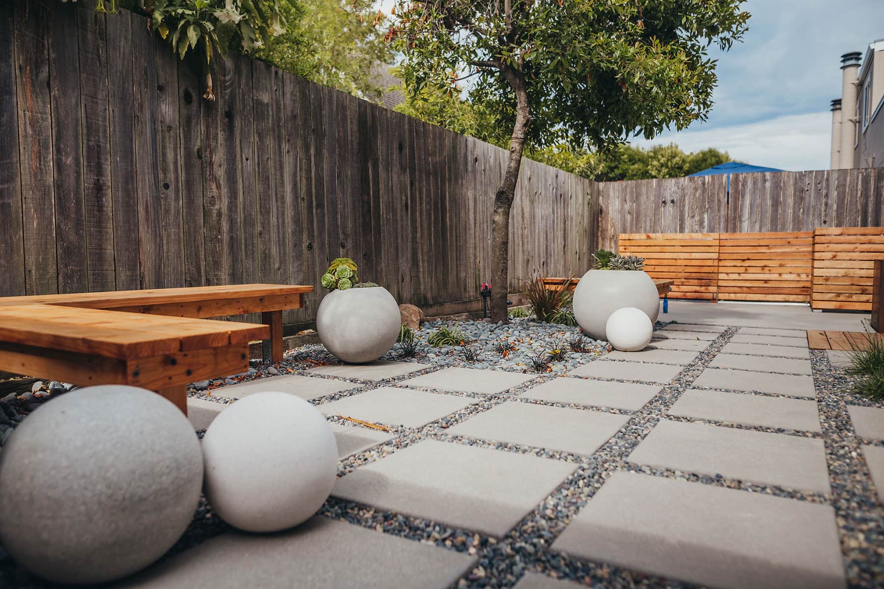 Backyard landscape featuring redwood fencing, wooden benches, white sphere planters in gravel beds, concrete and gravel patio pattern, and narrow redwood screening by K&D Landscaping