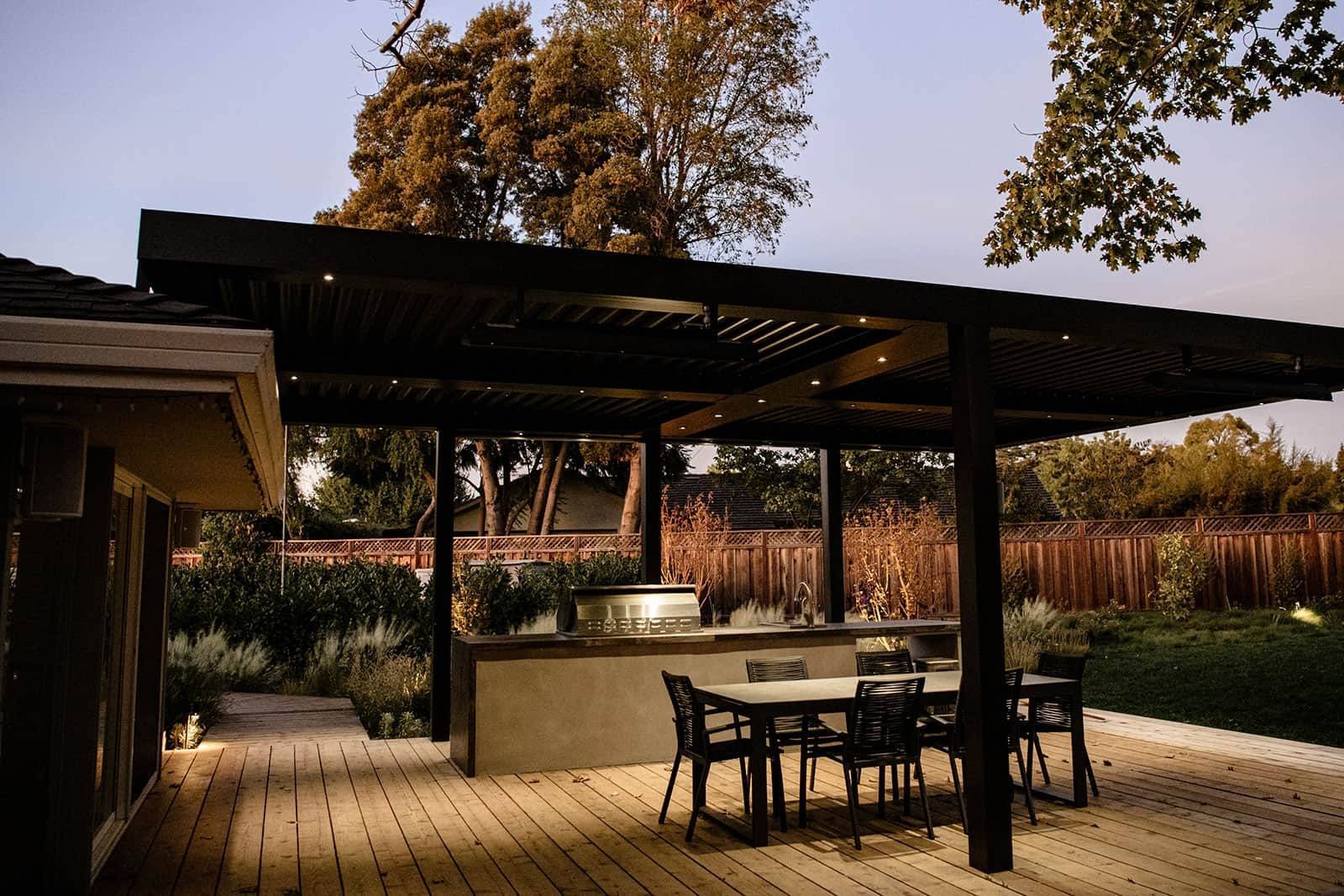 Evening outdoor kitchen and entertaining pergola with grill counter, dining table, wood deck, and landscape beds by K&D Landscaping