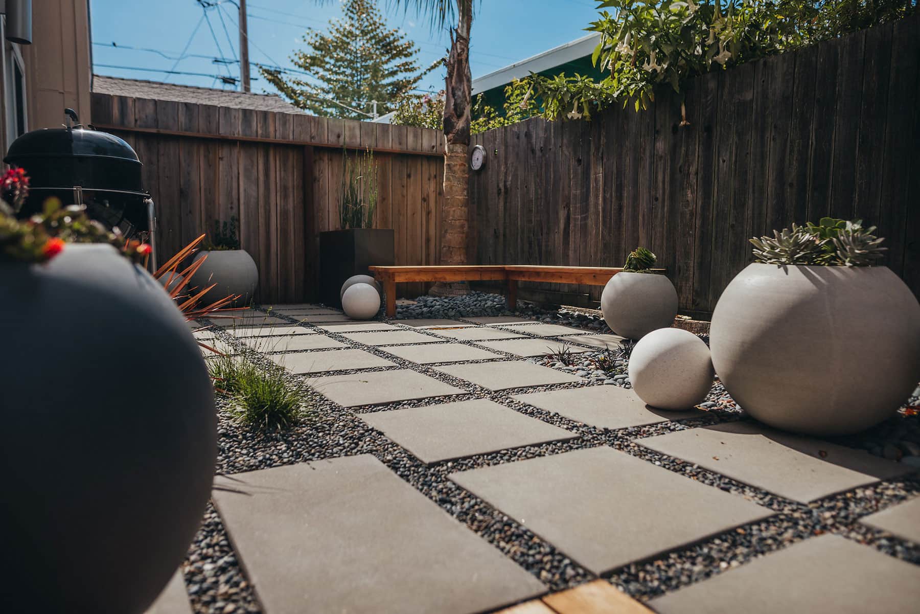 Full backyard view with redwood seating, white sphere planters featuring succulents and ornamental plants, concrete paver grid with gravel joints, and mature tree canopy by K&D Landscaping