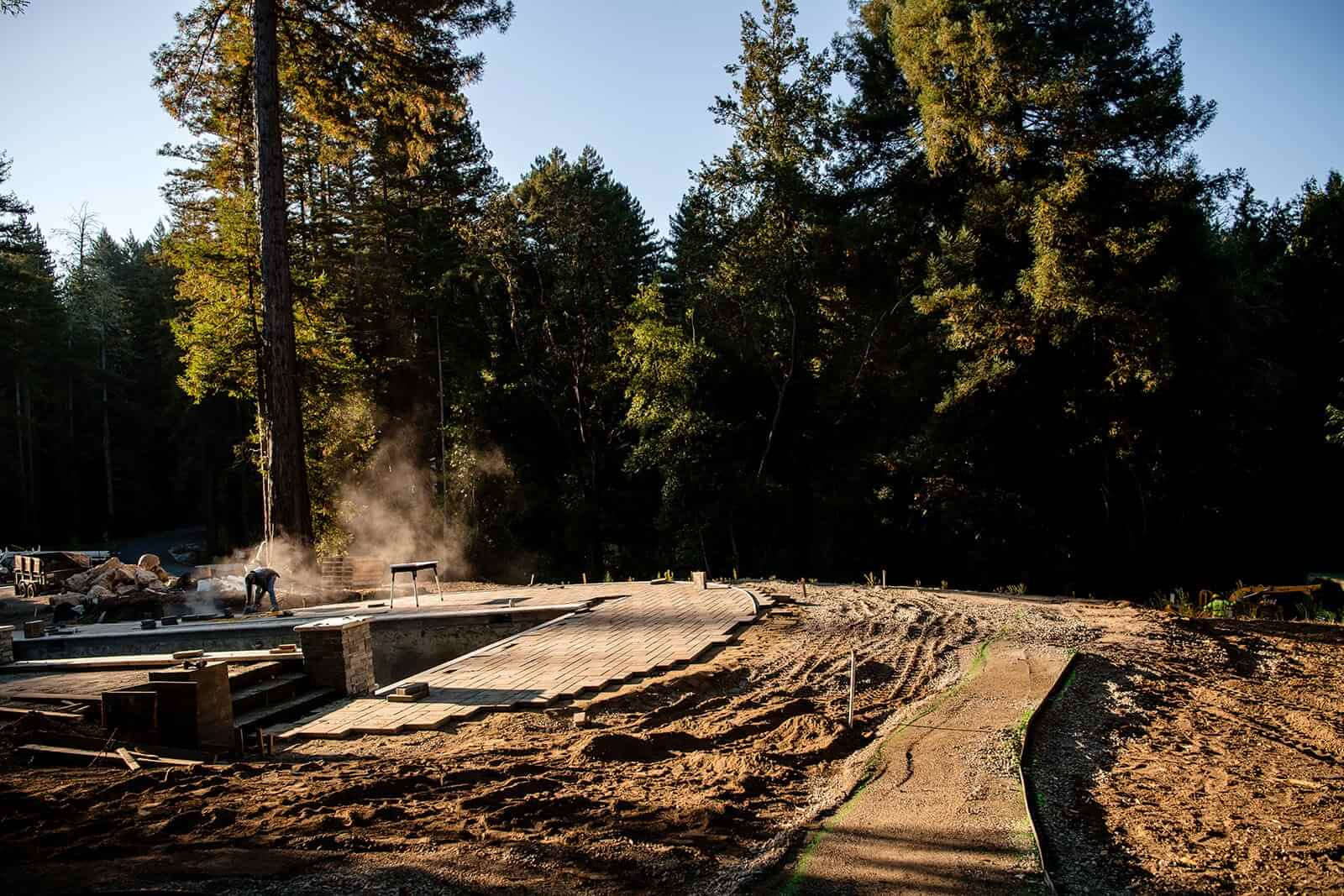 K&D Landscaping backyard hardscape under construction with newly laid pavers and graded soil among redwoods
