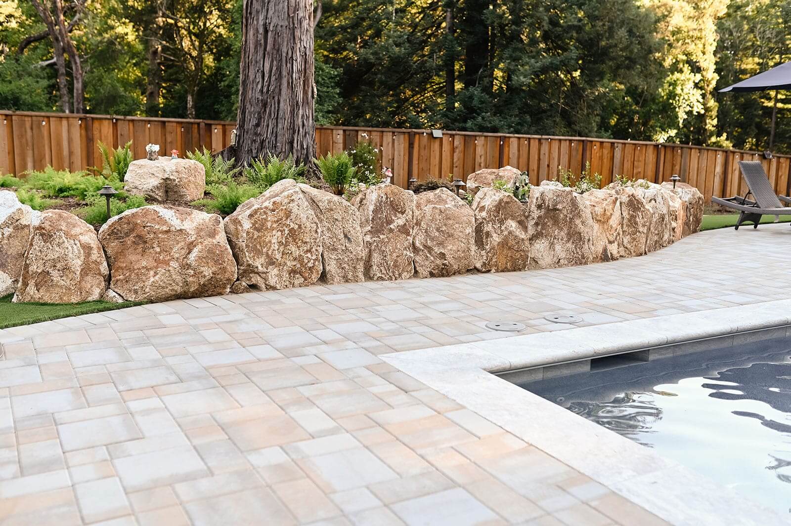 K&D Landscaping boulder retaining wall separating a fern planting bed from a paver pool deck