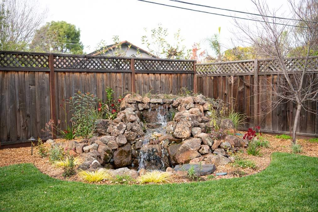 K&D Landscaping boulder waterfall water feature set into a mulch bed beside a manicured lawn