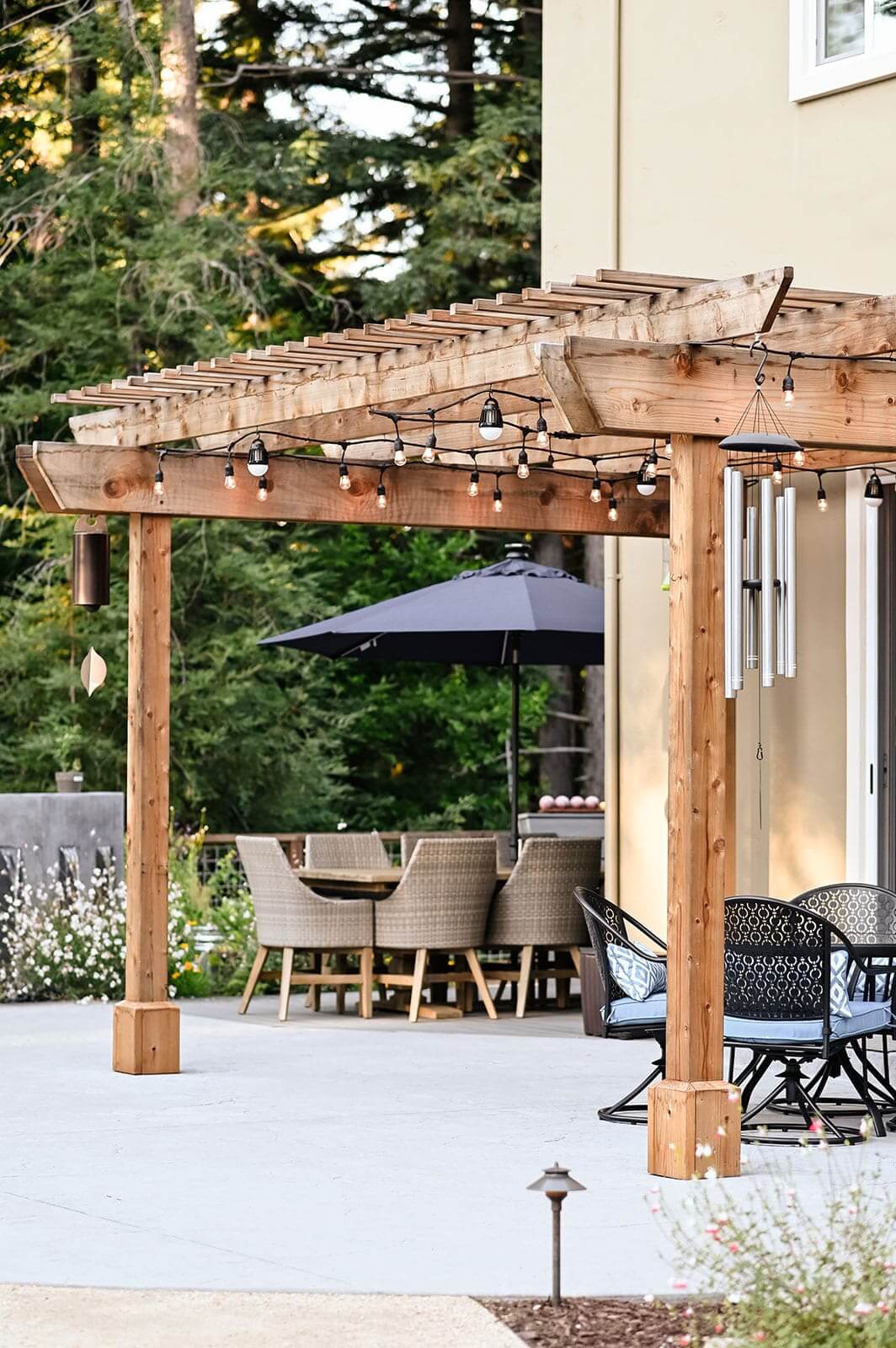 K&D Landscaping cedar pergola hung with string lights and wind chimes over a concrete patio dining area