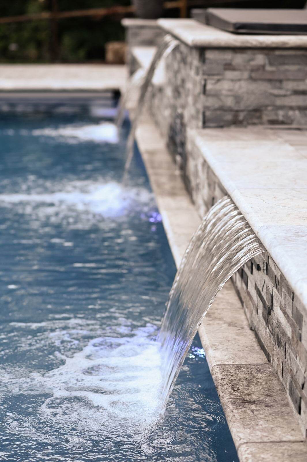 K&D Landscaping close-up of sheet waterfalls spilling over a travertine-capped stone wall into the pool