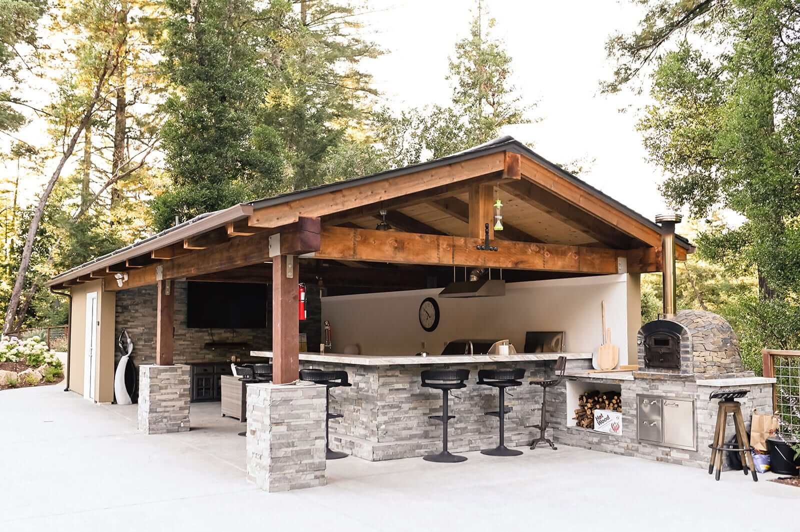 K&D Landscaping covered outdoor kitchen and bar pavilion with a stacked-stone counter and wood-fired pizza oven