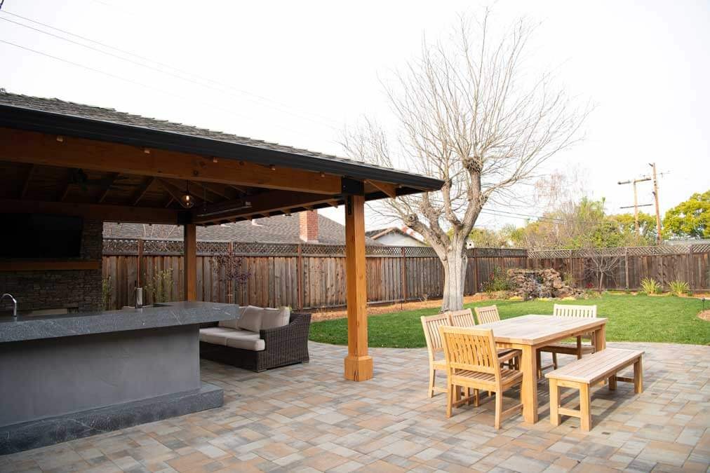 K&D Landscaping covered outdoor kitchen and lounge pavilion beside a teak dining set on a paver patio