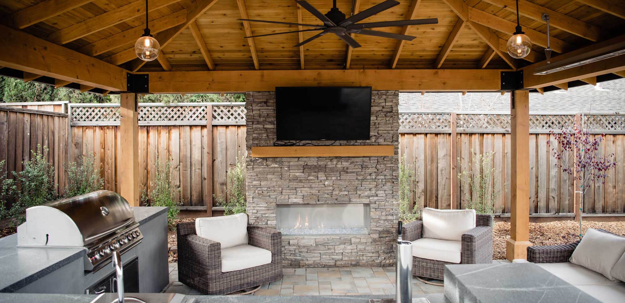 K&D Landscaping covered patio lounge with a stacked-stone fireplace, mounted TV and built-in grill