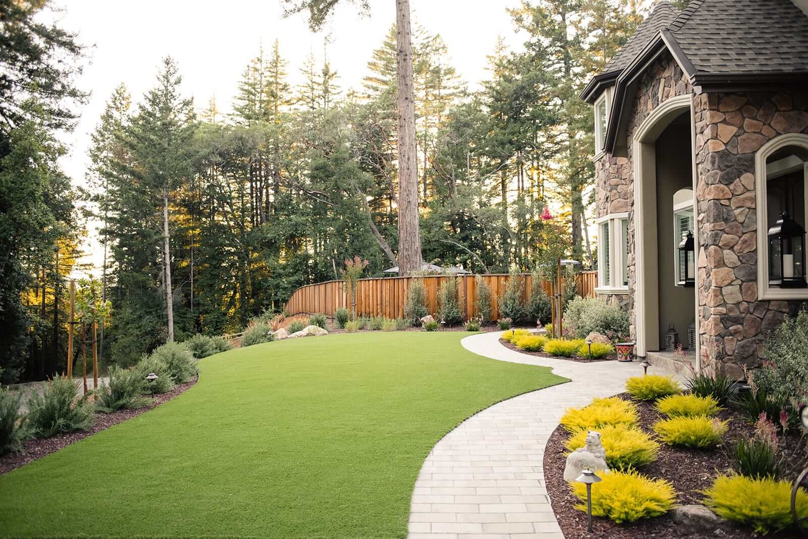 K&D Landscaping curved paver walkway and artificial turf lawn beside a stone-clad home with gold-mound shrubs