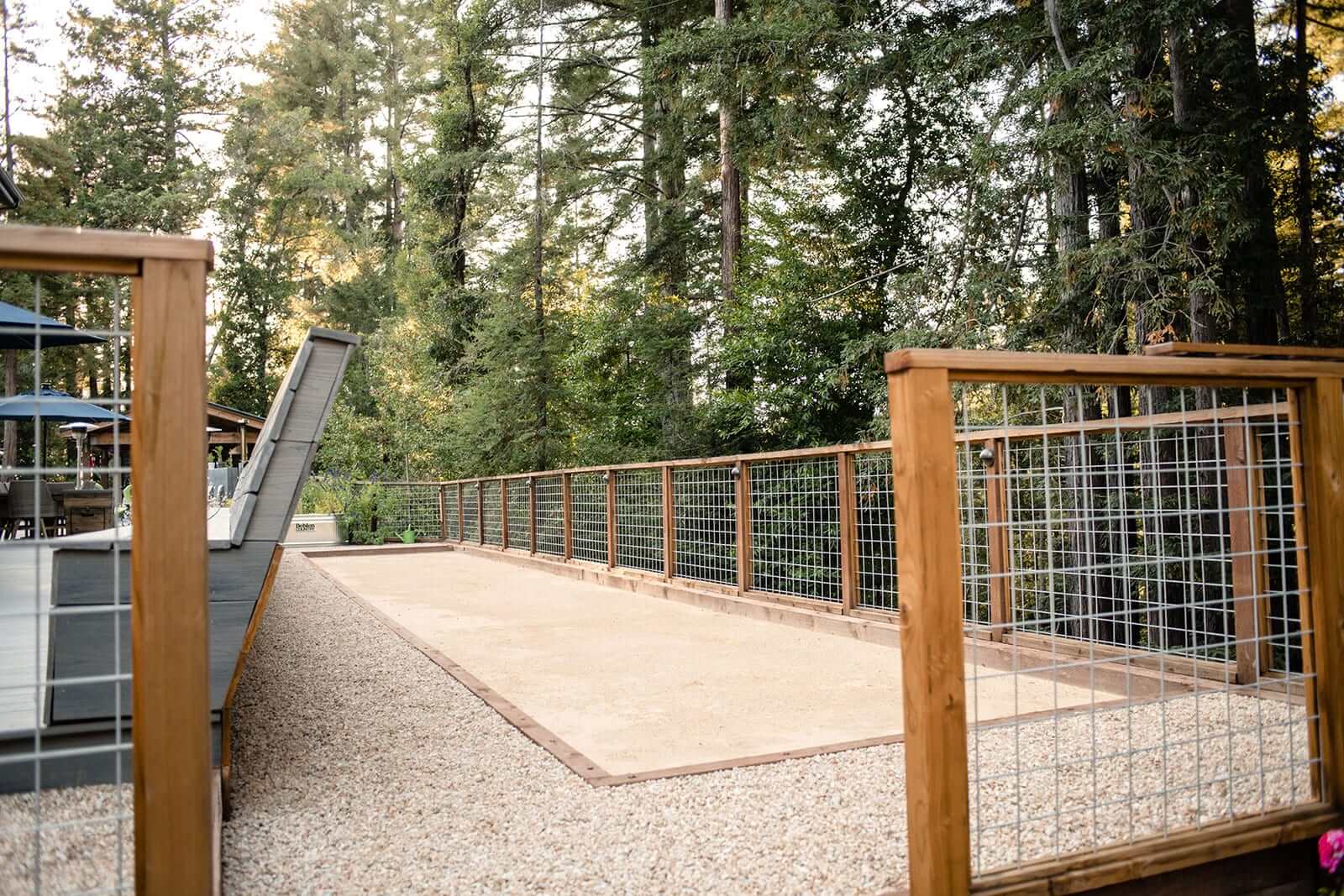K&D Landscaping decomposed-granite bocce court enclosed by a hog-wire fence among the redwoods