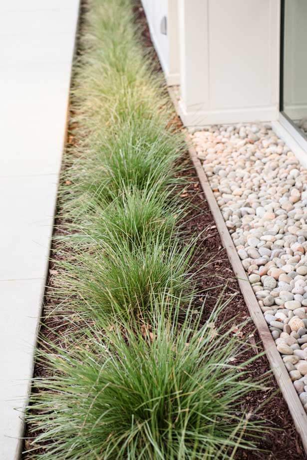 K&D Landscaping deer grass tufts in a mulch strip beside a river-rock drainage bed and walkway