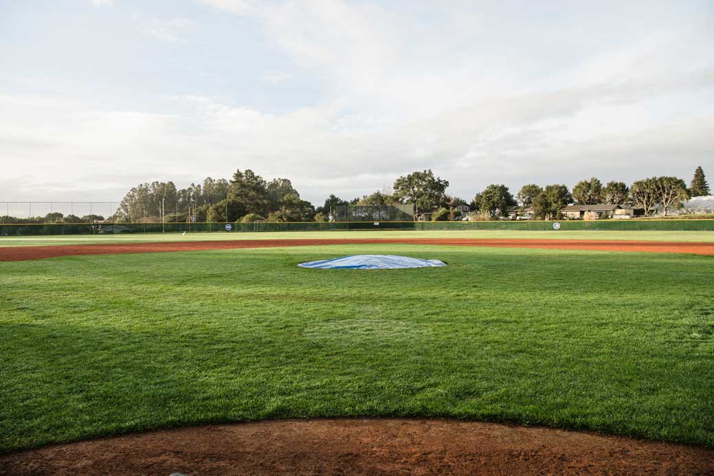 K&D Landscaping grass baseball infield with a tarp-covered pitching mound and outfield fence