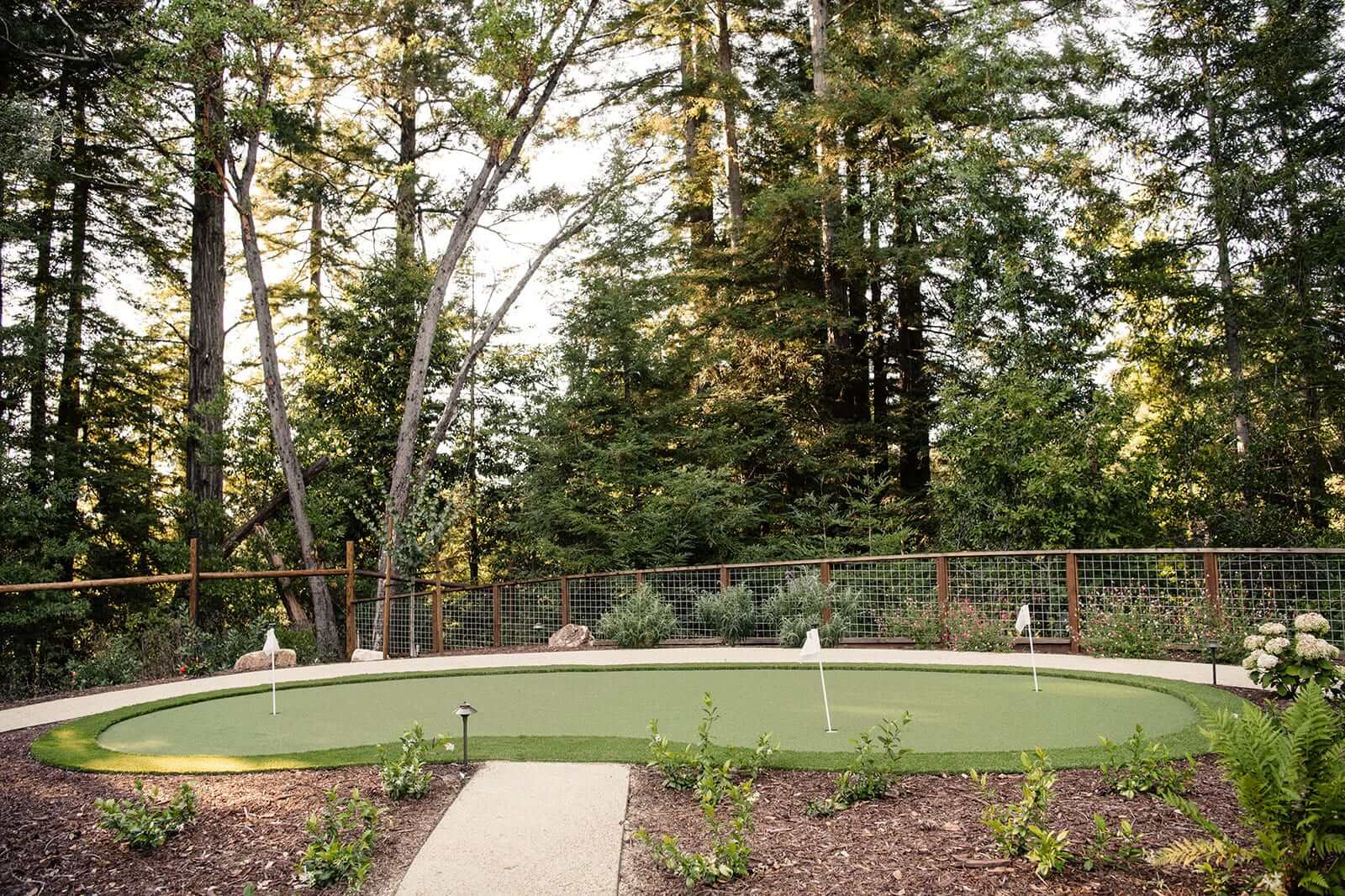 K&D Landscaping oval artificial turf putting green bordered by a welded-wire ranch fence and redwoods