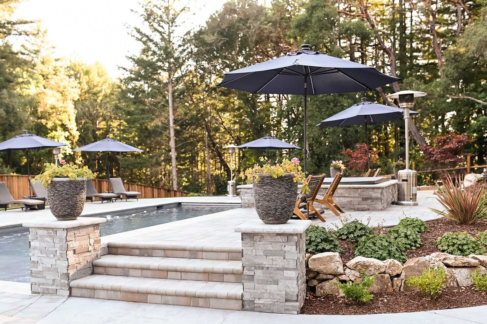 K&D Landscaping paver pool deck with stacked-stone steps and woven planters shaded by navy umbrellas