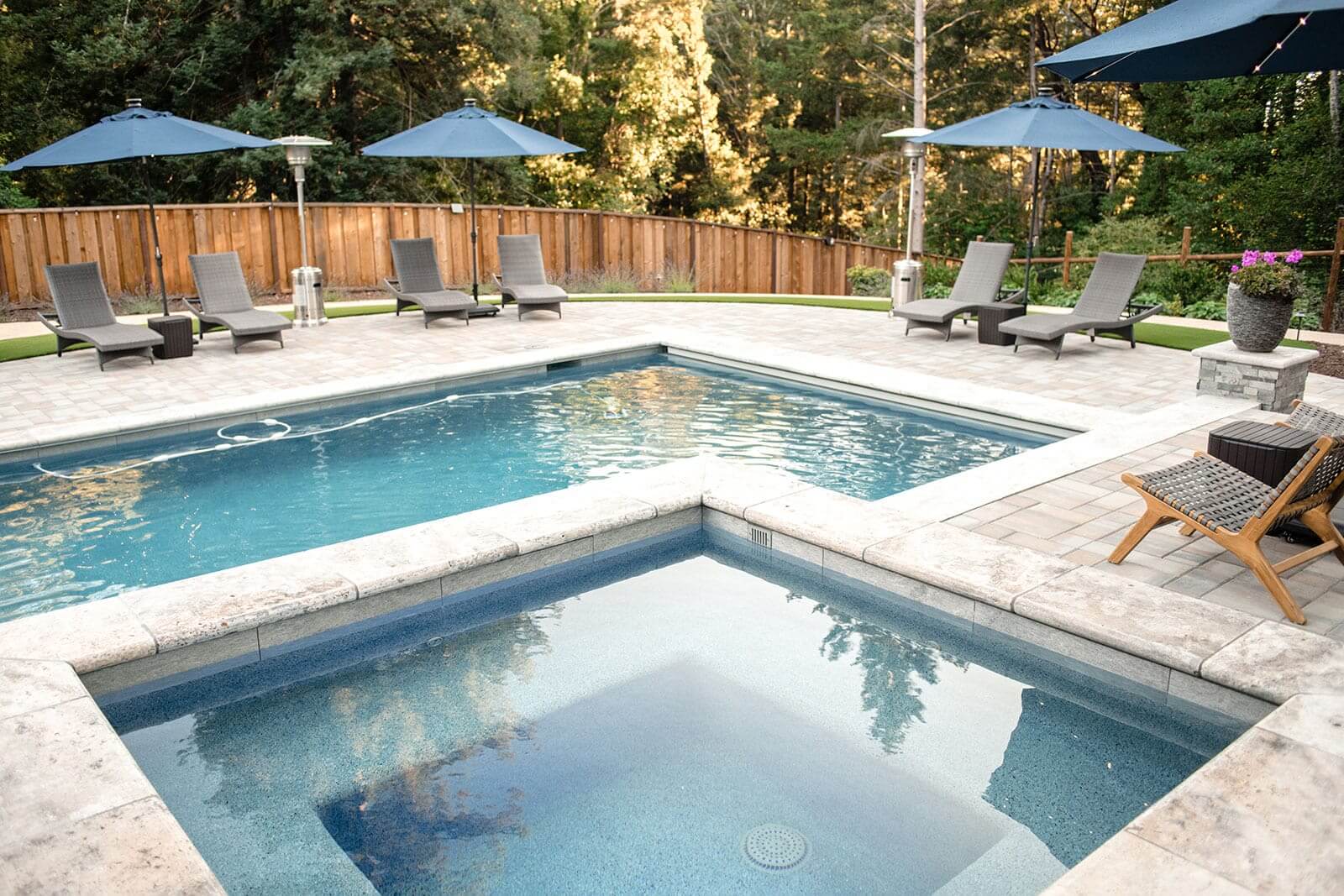 K&D Landscaping pool and raised spa with travertine coping framed by loungers and blue umbrellas