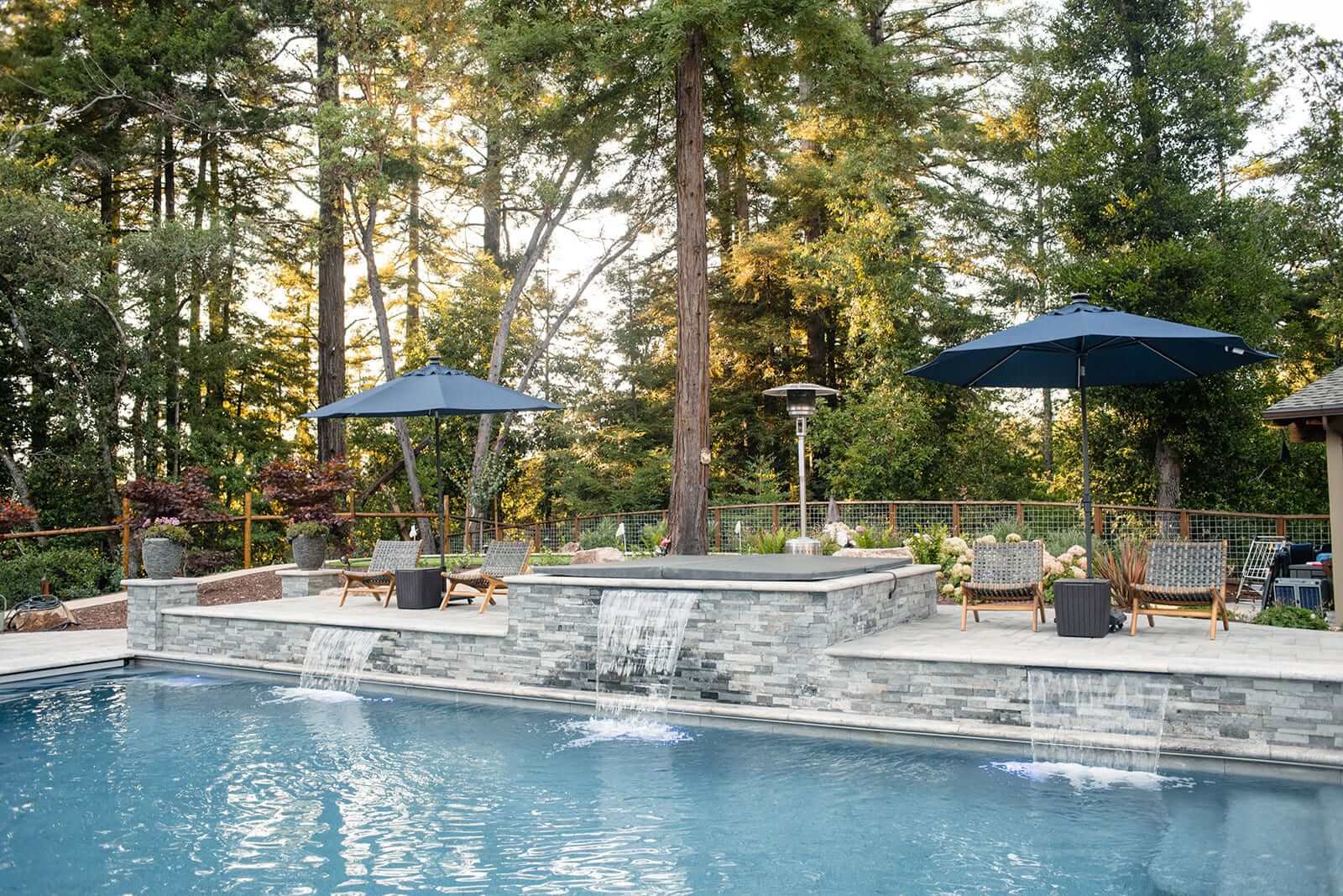 K&D Landscaping raised spa spilling sheet waterfalls into the pool with loungers and blue umbrellas