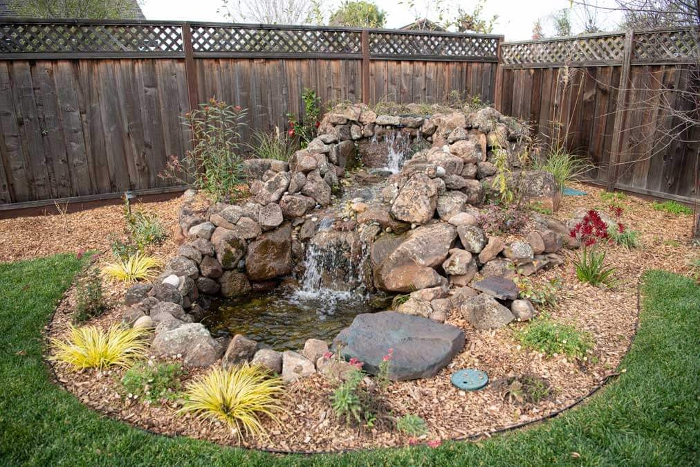 K&D Landscaping rock waterfall and pond framed by golden grasses and a mulched planting bed