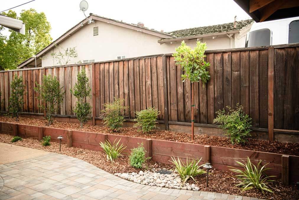 K&D Landscaping wood-tie raised planter bed along a fence with shrubs, river rock and path lighting