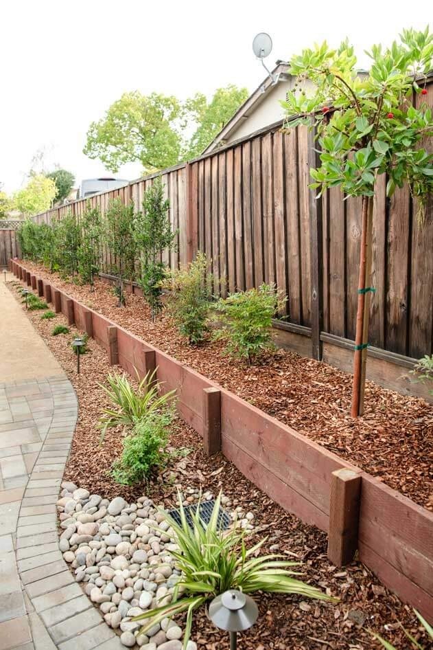 K&D Landscaping wood-tie raised planter bed with river rock and landscape lighting along a fence