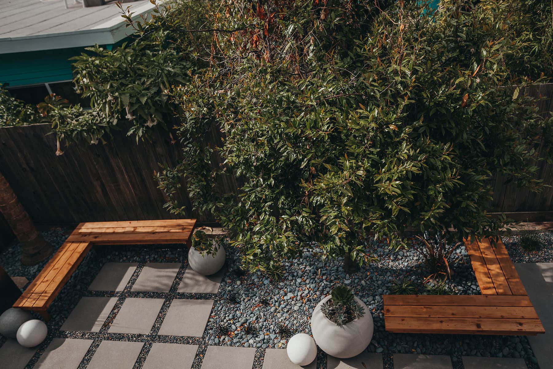 Modern residential backyard with concrete tile and gravel patio, redwood benches, white sphere planters with succulents, and ornamental shrub landscaping by K&D Landscaping