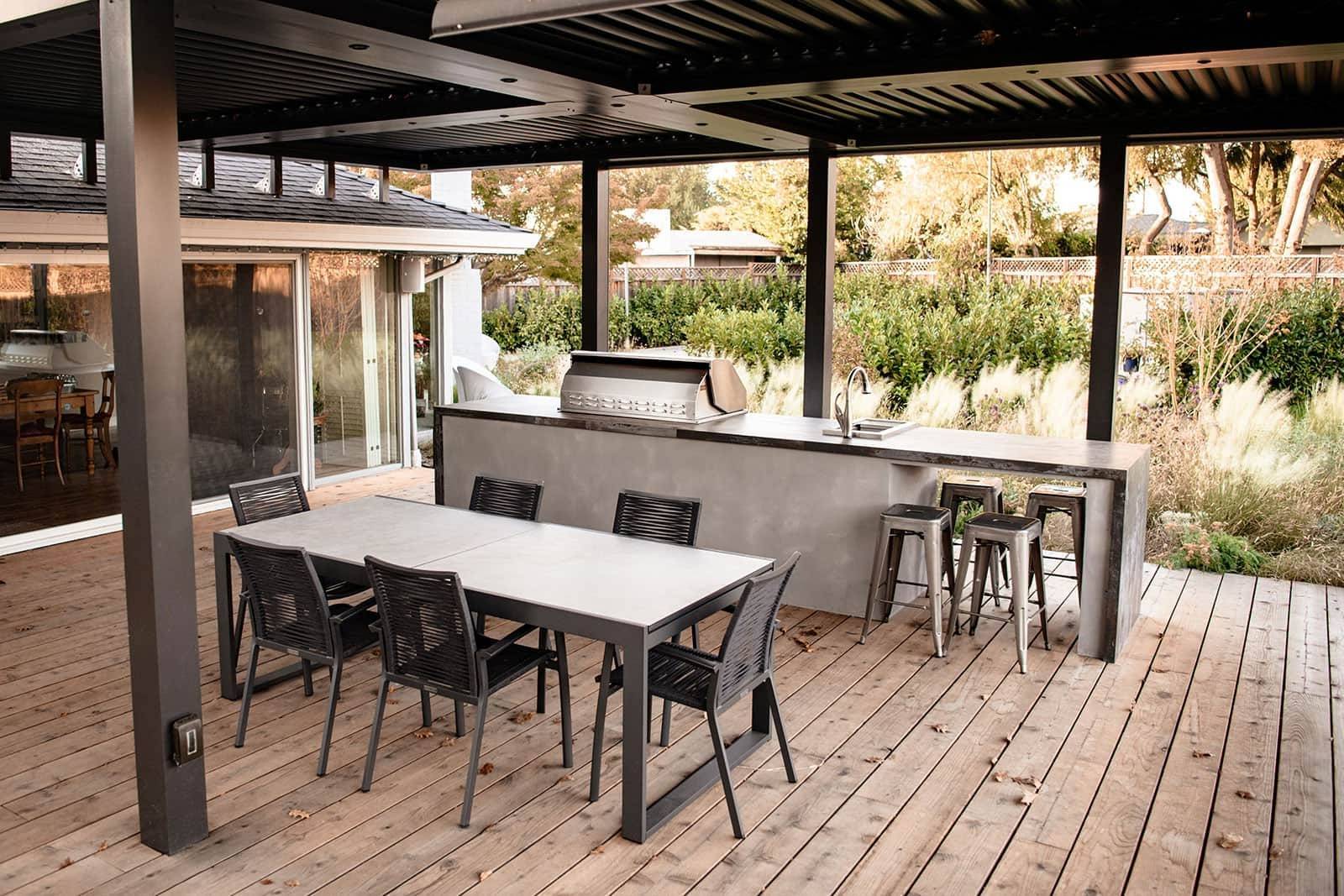 Pergola-covered outdoor kitchen and bar with dining table, grill station, wood decking, and garden plantings by K&D Landscaping