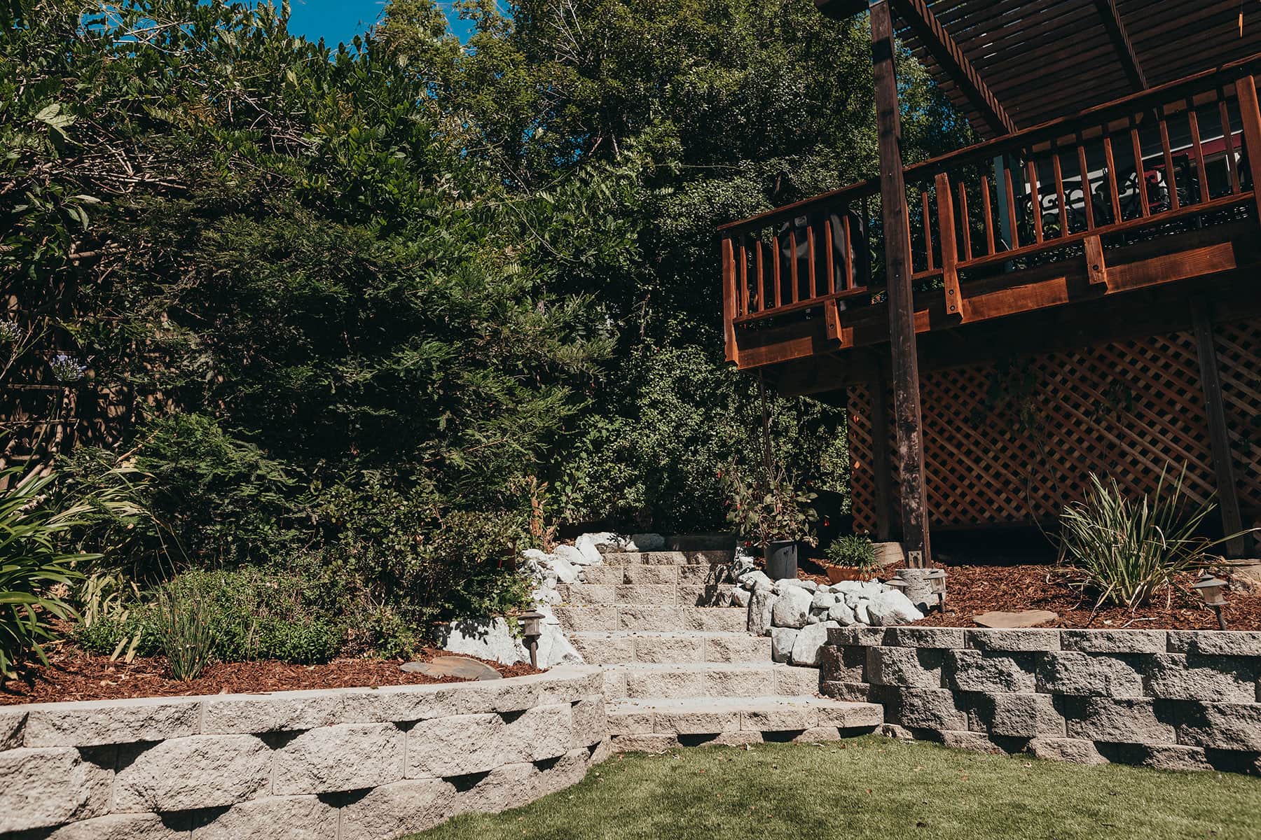 Redwood deck with natural stone retaining wall, gravel pathway, landscaping planting, and forest setting by K&D Landscaping 2