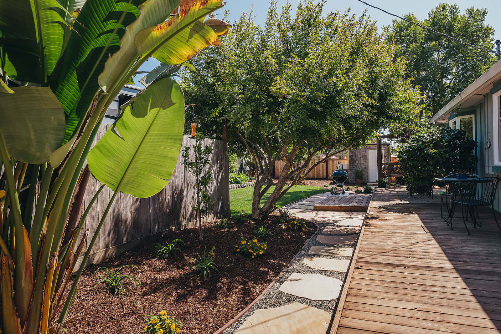 Residential backyard landscape with mature trees, tropical plantings including bananas, gravel beds, stone pathways, and modern hardscape by K&D Landscaping
