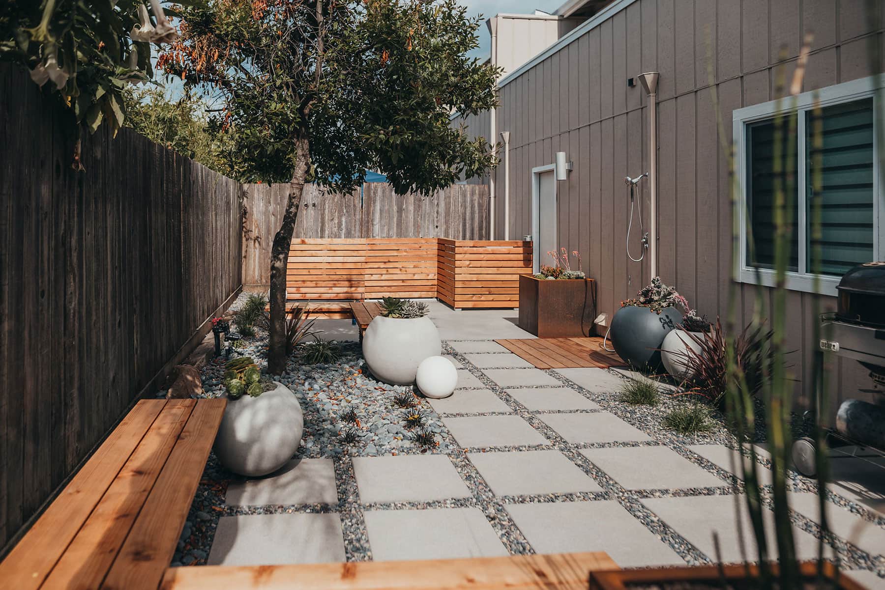 Residential yard renovation with redwood bench seating, white sphere planters, concrete paver grid, mature tree canopy, outdoor shower fixture, and modern landscape design by K&D Landscaping