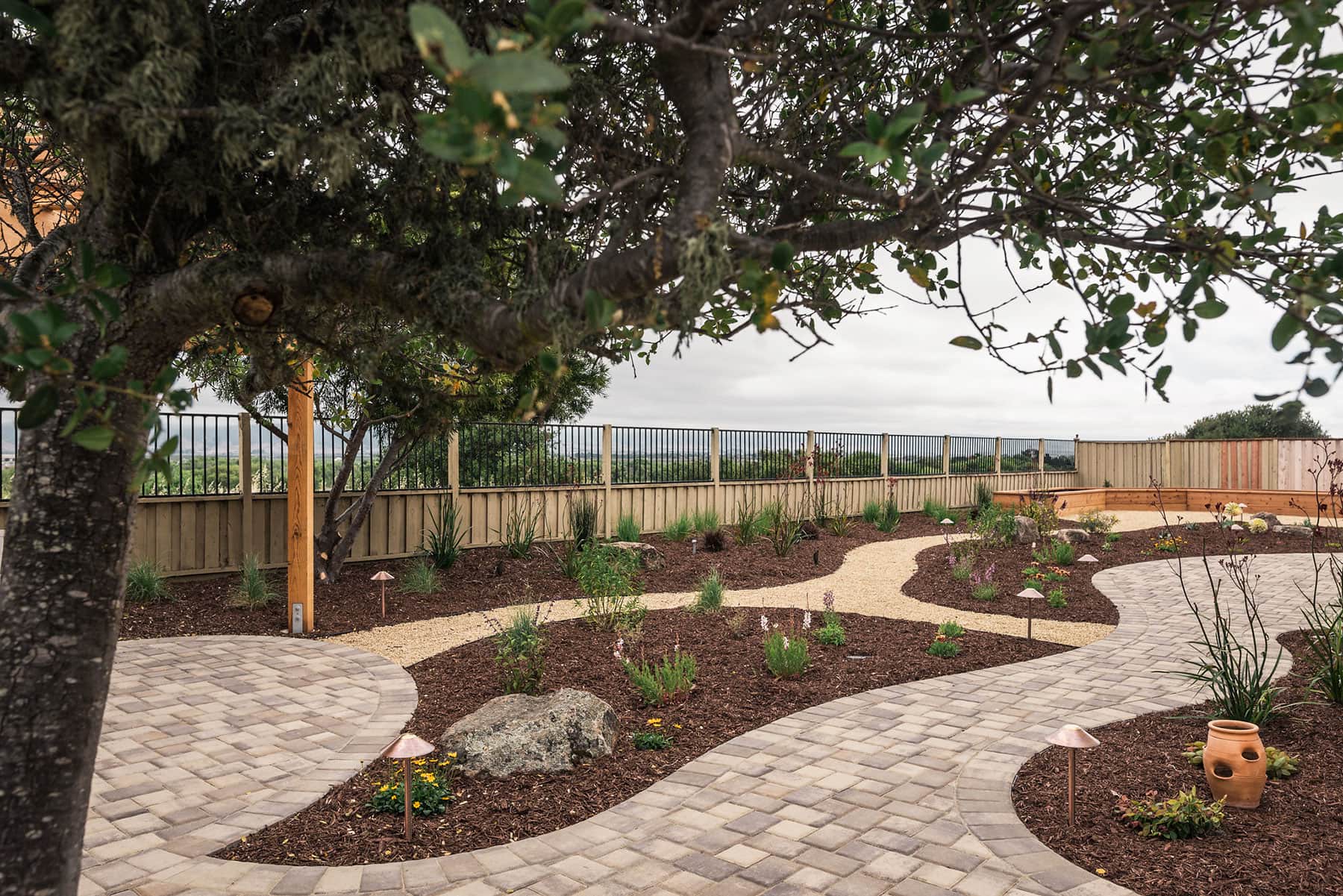 Wide backyard landscape with mature tree canopy, gravel and paver walkways, mulch planting beds, ornamental shrubs, decorative boulders, and modern design installed by K&D Landscaping
